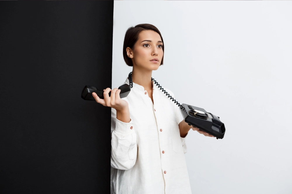 Eine Frau steht mit fragendem Blick und einem Telefon über den Schultern vor einer weißen Wand