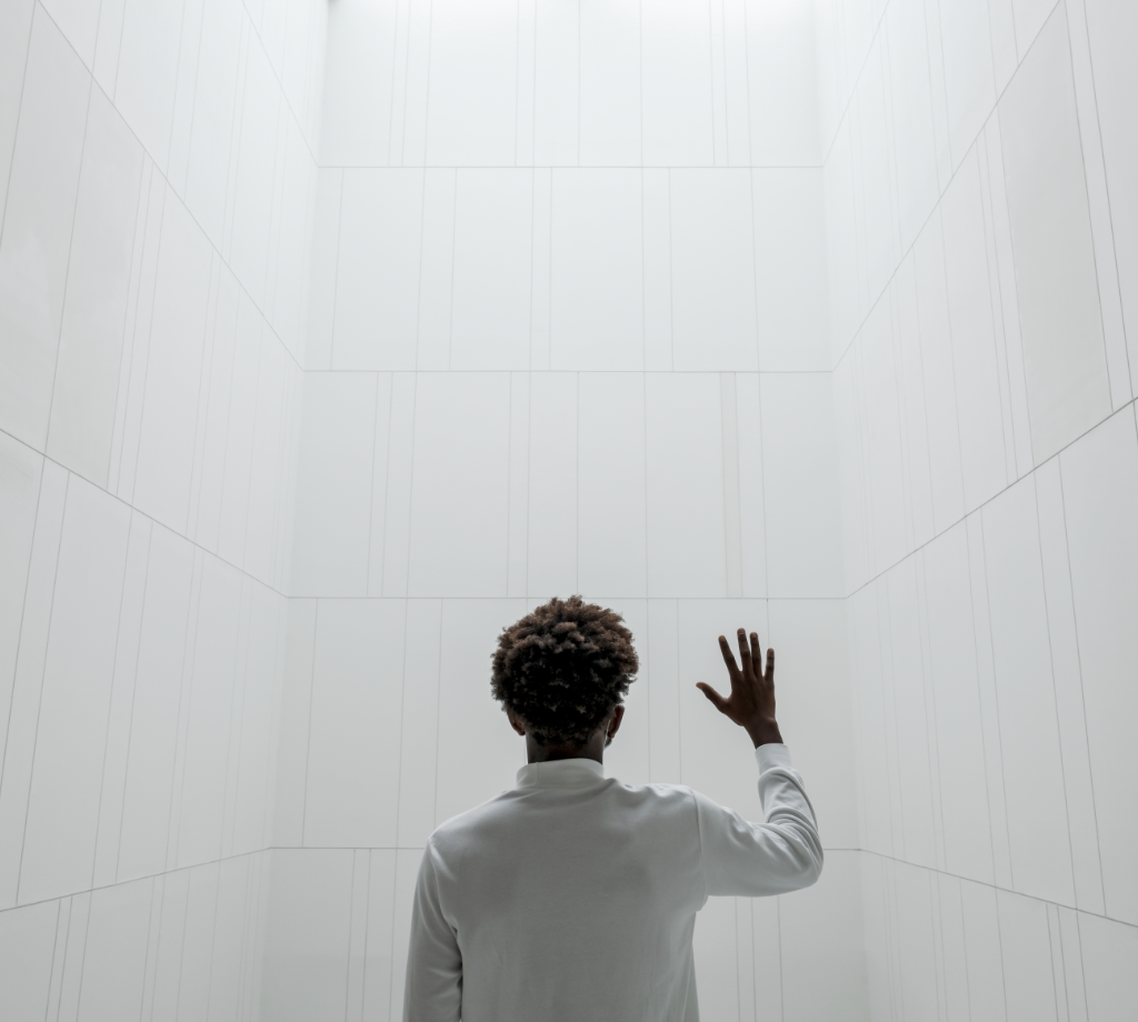 Ein Mann steht mit dem Rücken zur Kamera vor einer weißen Wand und hebt die Hand gestalterisch