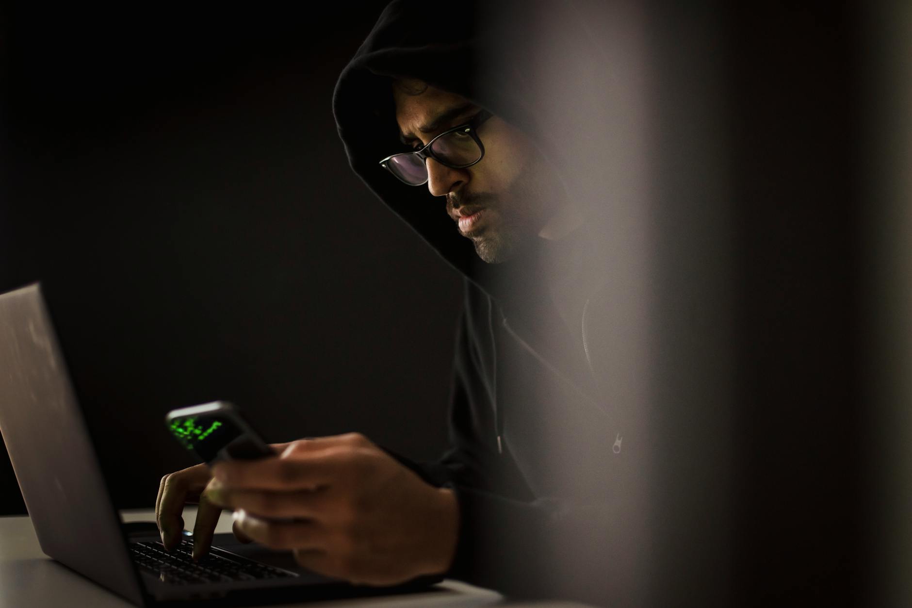 Crop bearded ethnic male computer geek in hoodie chatting on cellphone while typing on netbook in dark room