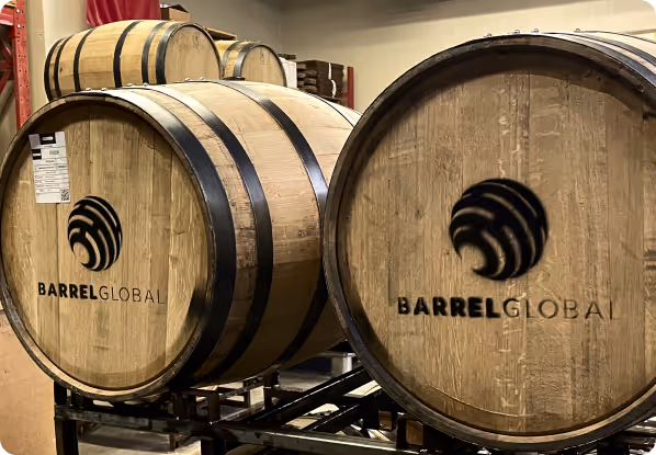 Wooden barrels with metal bands stacked on a rack, both labeled with the Barrel Global logo.