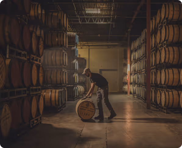 Man in a warehouse rolling a wooden barrel between large stacks of barrels on metal racks.
