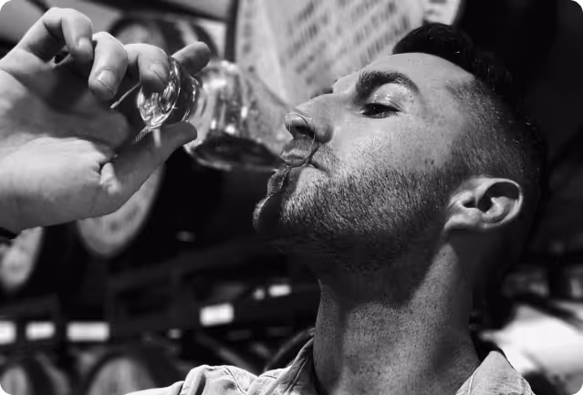 Man tasting liquor or whiskey from a small glass, with barrels in the background.