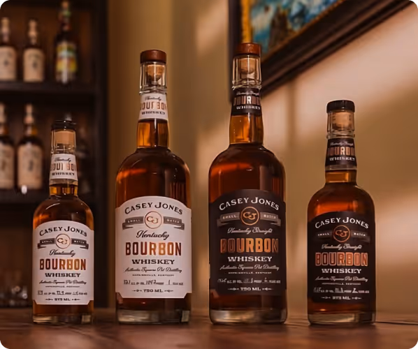 Four bottles of Casey Jones Kentucky Bourbon Whiskey in varying sizes displayed on a wooden surface with a blurred background featuring shelves and a framed picture.