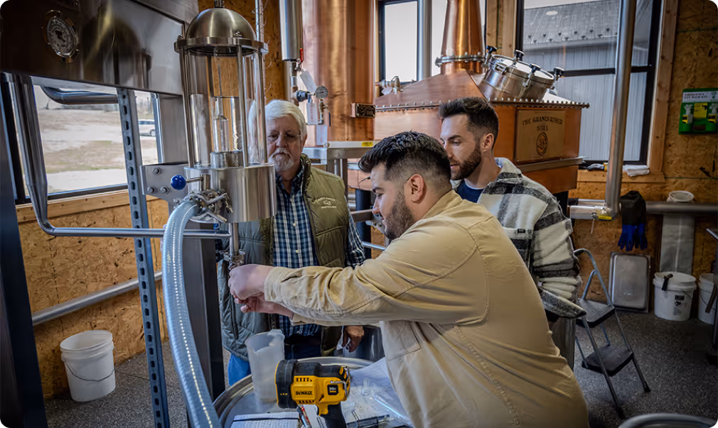 Three men working together at a distillery still, inspecting equipment and taking notes.