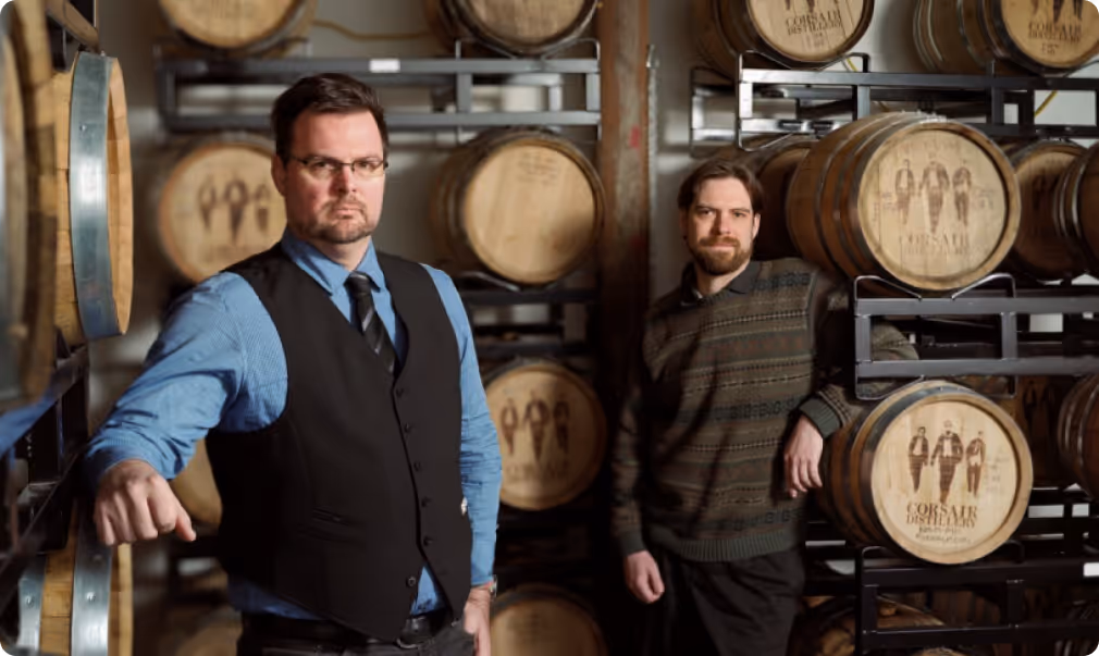 Two men standing among stacked wooden barrels in a distillery, one wearing a blue shirt with a black vest and tie, the other in a patterned sweater.