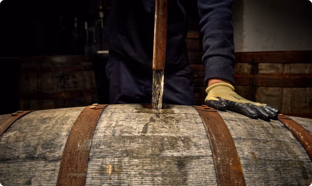Worker wearing gloves extracts liquid from a wooden barrel with a metal spout in a dimly lit cellar.