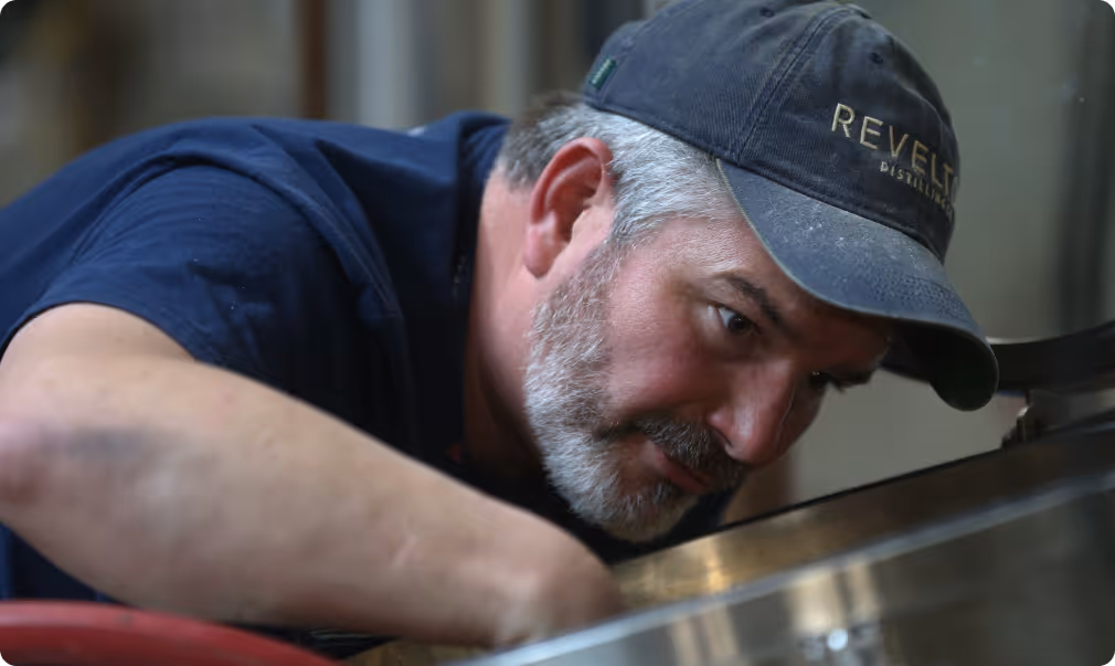 Man with gray beard wearing a dark cap and navy shirt closely inspecting a large metal container.