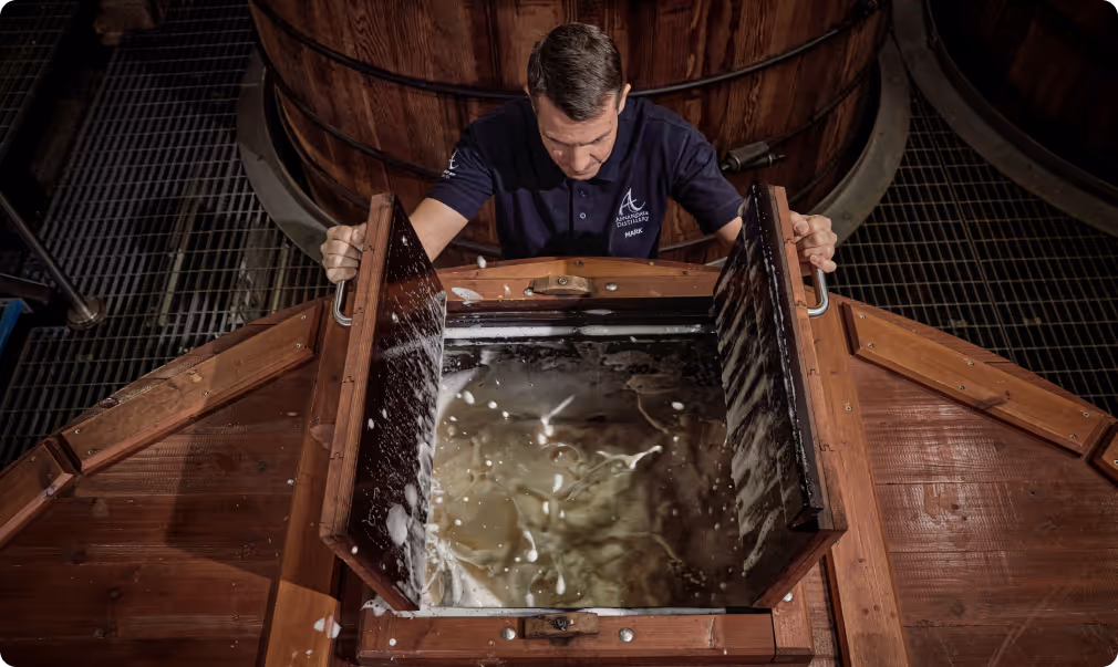 Man in navy shirt opening a wooden hatch revealing a frothy liquid inside a large wooden vat in a distillery.