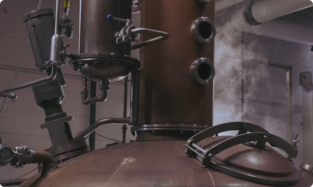 Close-up of industrial distillation or brewing equipment with steam rising in a factory setting.