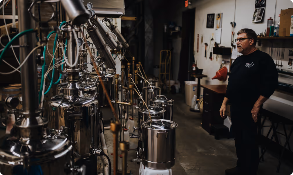 Man standing in a distillery workshop with multiple stainless steel stills and pipes.