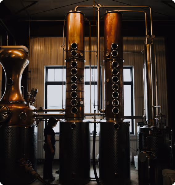 Person standing near large copper distillation columns in an industrial distillery with metal tanks and pipes.