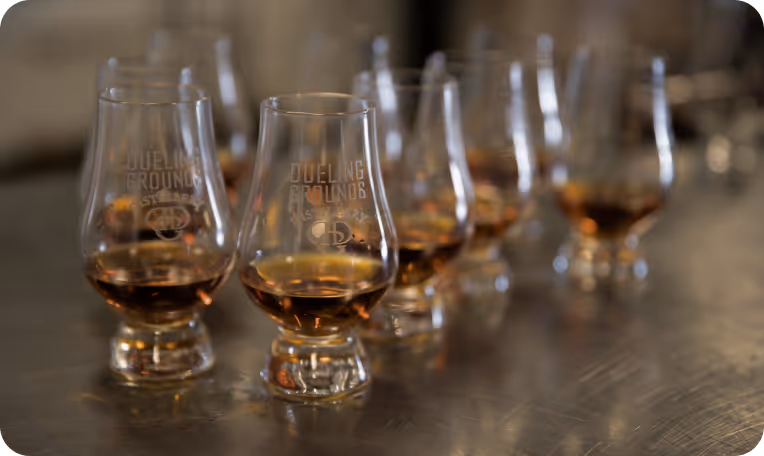 Seven tasting glasses with amber liquid arranged in a staggered row on a reflective surface.