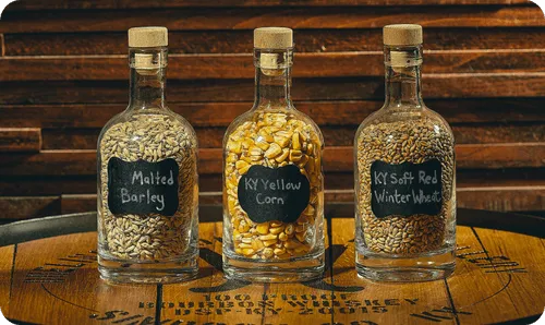 Three glass bottles with cork stoppers, filled with malted barley, yellow corn, and soft red winter wheat, placed on a wooden barrel top.