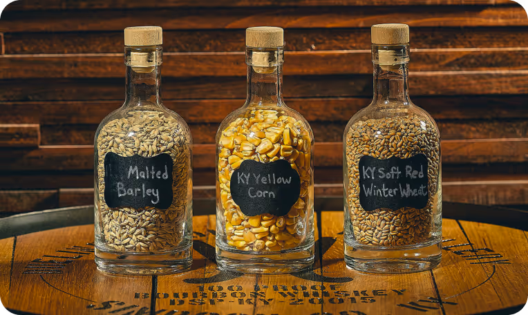 Three glass bottles with cork stoppers, filled with malted barley, yellow corn, and soft red winter wheat, placed on a wooden barrel top.