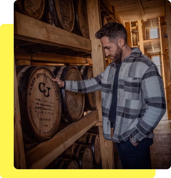 Man in a checkered jacket inspecting barrels of Casey Jones Distillery in a wooden storage room.
