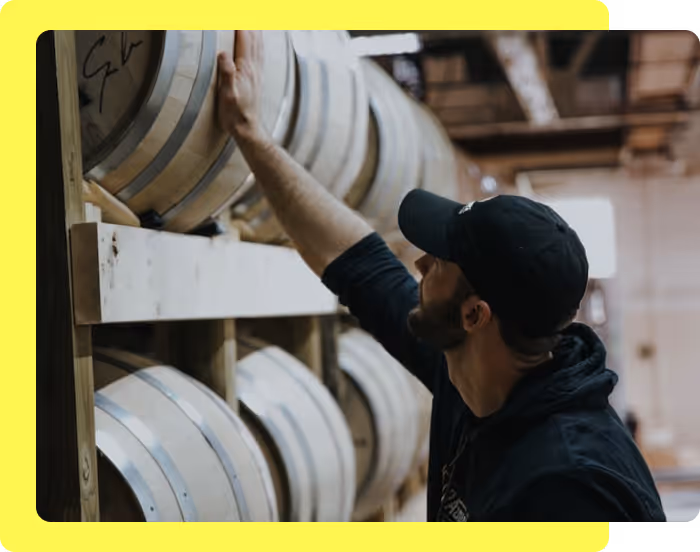 Man in black cap and hoodie inspecting wooden barrels stacked on shelves in a warehouse.