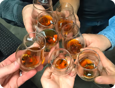 Seven people holding small glasses with amber-colored whiskey, toasting together.