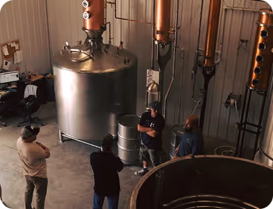 Four men standing and talking inside an industrial distillery with large stainless steel tanks and copper pipes.