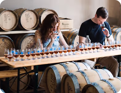 Two people tasting whiskey samples at a table in a barrel storage room.
