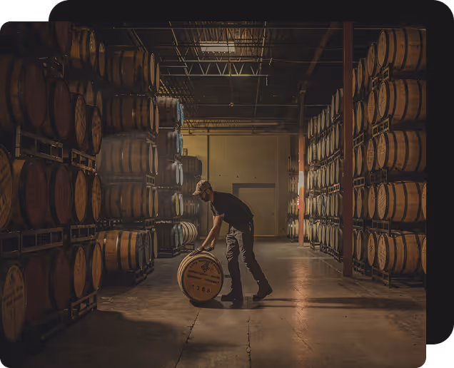 Person rolling a wooden barrel inside a warehouse filled with stacked barrels.
