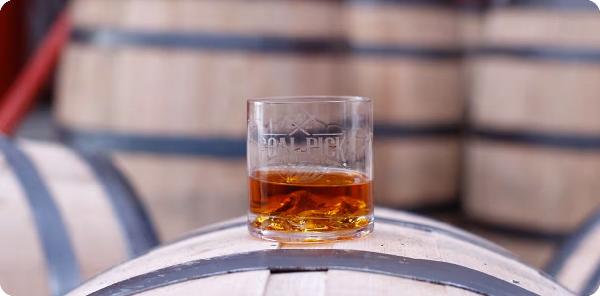 Glass of whiskey with ice placed on top of a wooden barrel in a distillery.