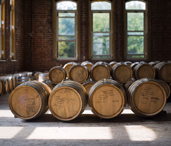 Rows of wooden barrels in a dimly lit room with large windows and brick walls, likely a distillery storage area.