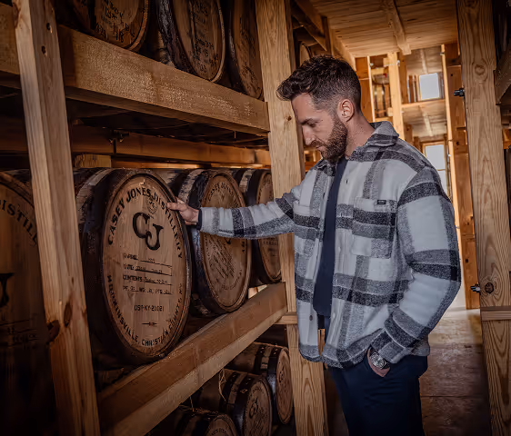 Man in a checkered jacket inspecting wooden barrels in a dimly lit storage room.