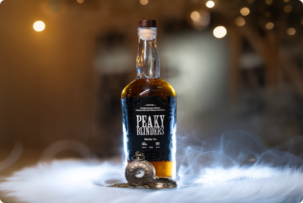 Bottle of Peaky Blinders whiskey on a surface with smoke and a vintage pocket watch in front, against a blurred background with warm lights.
