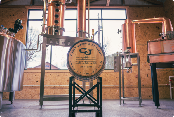 Copper distillation equipment and a wooden barrel labeled Casey Jones Distillery in a rustic distillery room with large windows.