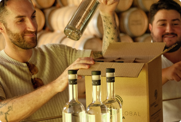 Two men packing clear glass bottles into a cardboard box in a room filled with wooden barrels.
