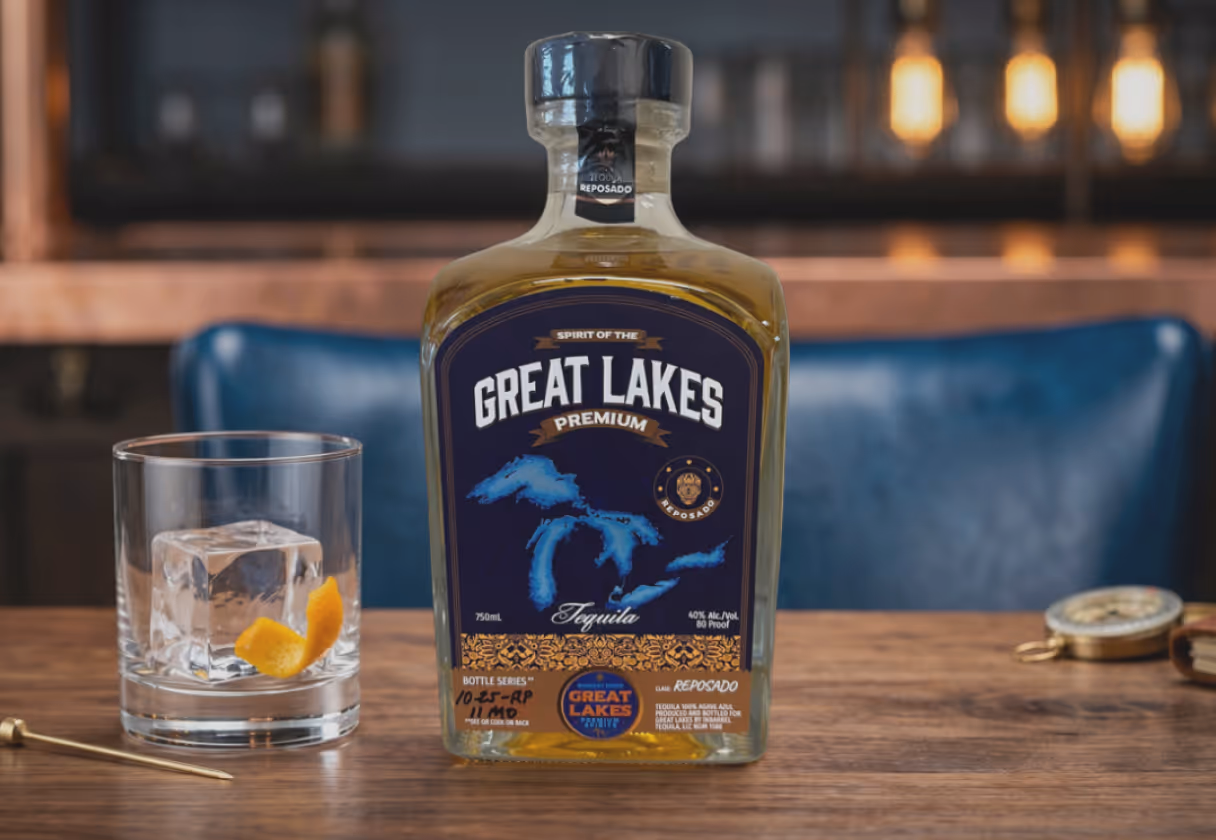 Bottle of Great Lakes Premium Reposado Tequila beside a glass with an ice cube and orange peel on a wooden table.