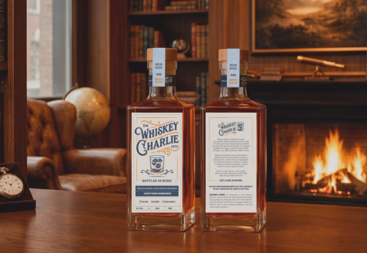 Two bottles of Whiskey Charlie Bourbon on a wooden table in a cozy room with a leather armchair, globe, and a lit fireplace.