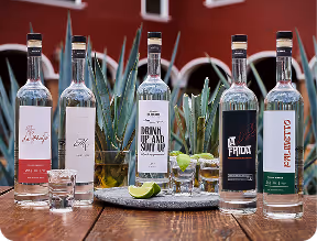 Six bottles of assorted alcoholic beverages with shot glasses and lime wedges on a wooden table outdoors.