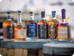 Six different bottles of whiskey lined up on a wooden barrel with a blurred background.