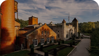 Historic castle-like building with towers, brick structures, and a pathway surrounded by greenery under a partly cloudy sky.