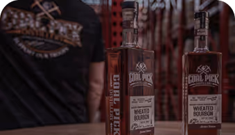 Two bottles of Coal Pick Distillery Wheated Bourbon on a table with a person in a black Coal Pick shirt.
