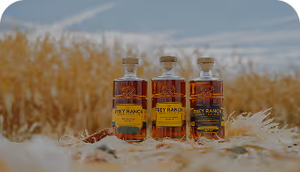 Three bottles of Frey Ranch whiskey on grass with a field and blue sky background.