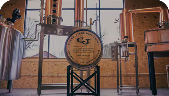 Copper distillation equipment with a labeled barrel in a light wooden room with windows.