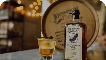 Bottle of Journeyman Distillery Last Feather Rye Whiskey with a poured glass on wood table.