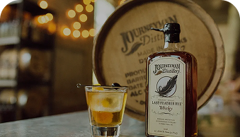 Bottle of Journeyman Distillery Last Feather Rye Whiskey with a poured glass on wood table.
