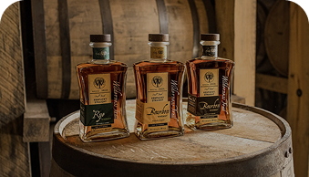 Three rectangular whiskey bottles on a wooden barrel in front of large wooden barrels.