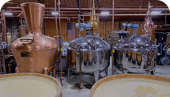 Copper and stainless steel distillation equipment inside a distillery with fermenting tanks in the foreground.