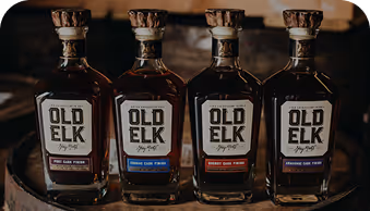 Four Old Elk whiskey bottles in a row with different barrel finish labels on a wooden surface.