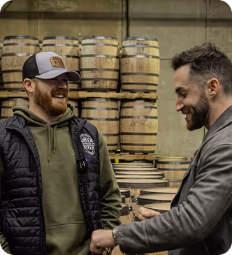 Two men smiling and chatting in a barrel storage room, one wearing a cap and vest, the other a blazer.