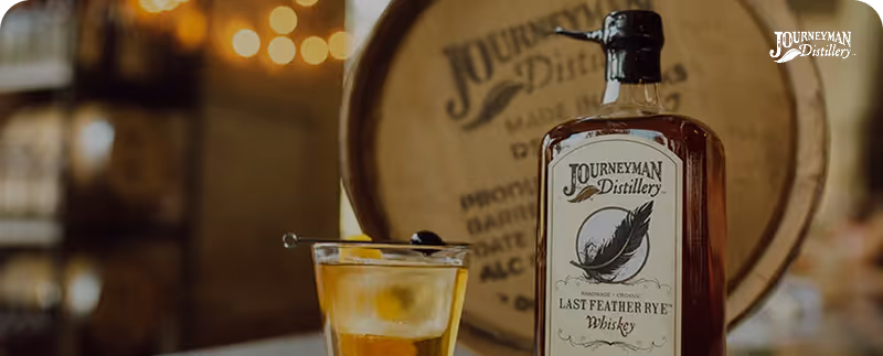 Bottle of Journeyman Distillery Last Feather Rye whiskey next to a glass with ice and a cocktail garnish.