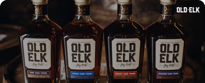 Four Old Elk whiskey bottles with different cask finishes lined up side by side on a wooden surface.
