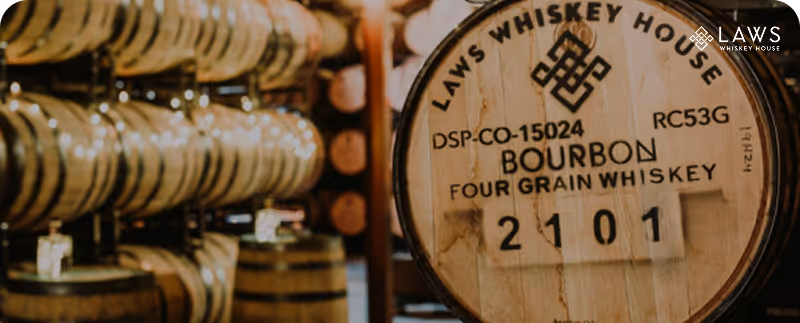 Wooden bourbon barrel labeled Laws Whiskey House Four Grain Whiskey in a warehouse with stacked barrels.