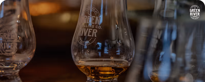 Glass with partial whiskey pour with Green River Distilling Company logo on a wooden surface.