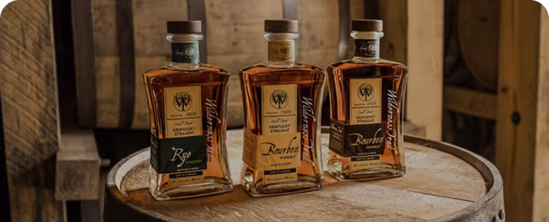 Three square bottles of Kentucky Straight Whiskey on a wooden barrel in a rustic cellar.