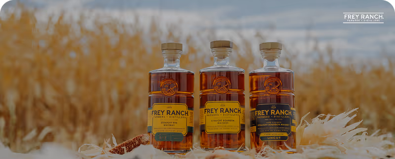 Three bottles of Frey Ranch whiskey arranged on straw with a field backdrop and Frey Ranch logo.
